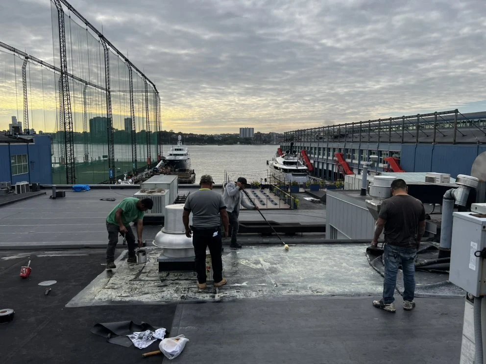 Roofing crew working on a flat commercial rooftop near the waterfront, applying materials and tools with boats and buildings in the background.