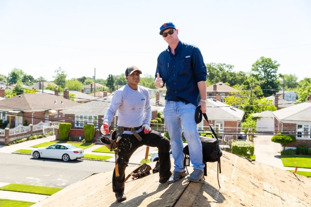 Two roofers standing on a residential rooftop, wearing work gear and tools, representing professional roofing services in the neighborhood.