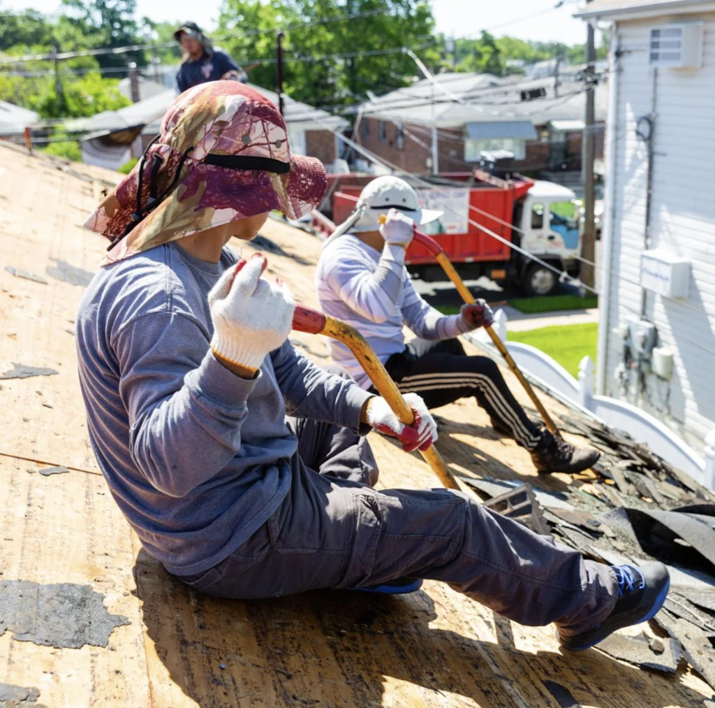 Roofers sitting on a residential rooftop while removing old shingles with hand tools during roof replacement work