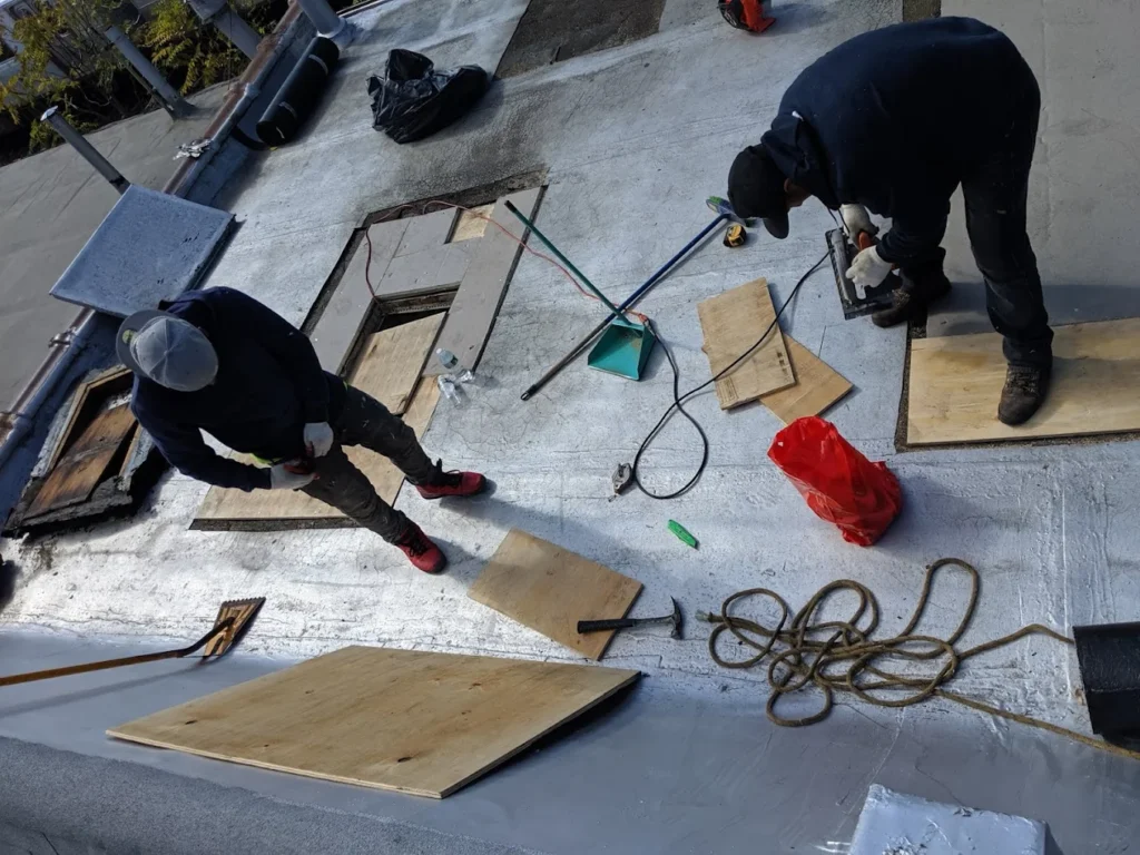 Two workers repairing a flat roof with plywood and tools, Manhattan roofing contractors installing new roof sections.